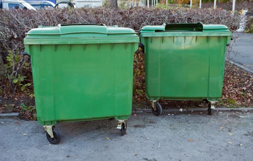 Two large green commercial wheelie bins with black plastic lids and small caster wheels are positioned side by side on a paved surface, with the bin on the left being slightly larger than the one on the right. The bins are made of durable plastic with a smooth finish and are situated outdoors, with a background of leafless shrubs and a metal fence indicating a typical urban or suburban street environment. The surrounding area appears clean and well-maintained, consistent with a commercial or municipal waste collection setting in Stratford. The image emphasizes the typical equipment used by rubbish collection services such as Commercial Waste Removal Stratford for handling general waste in local communities.