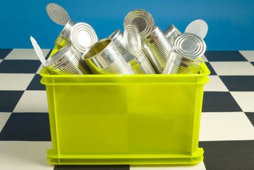 A bright yellow plastic container filled with numerous empty, silver metal cans, some with open tops, placed on a checkerboard black and white tiled floor against a plain blue wall background. The cans are stacked and leaning in various directions within the container, with some appearing crumpled or dented. There are no labels or markings on the cans. The scene suggests a waste collection or disposal environment, typical of rubbish removal services in Stratford, with the container ready for collection or recycling. The image is well-lit, emphasizing the glossy metallic surfaces of the cans and the vibrant yellow of the container, creating a clear and professional visual appropriate for a commercial waste removal context.