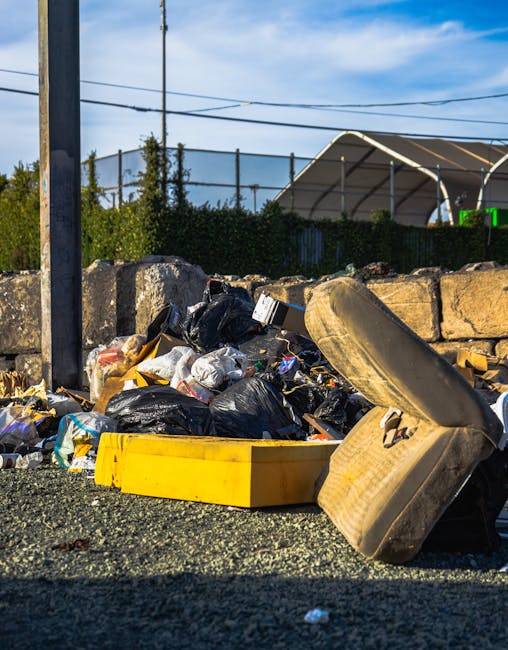 A narrow urban alleyway filled with piled waste and debris, with various discarded cardboard boxes, plastic bags, and other rubbish stacked at the far end. In the foreground, a large, grey, weathered 
