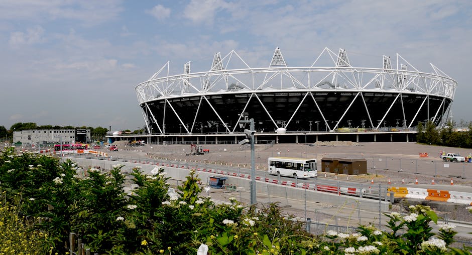 A wide-angle view of the interior of a large, modern stadium with a curved, partially open roof structure made of metal and glass panels. The underside of the roof displays a grid-like pattern with vi