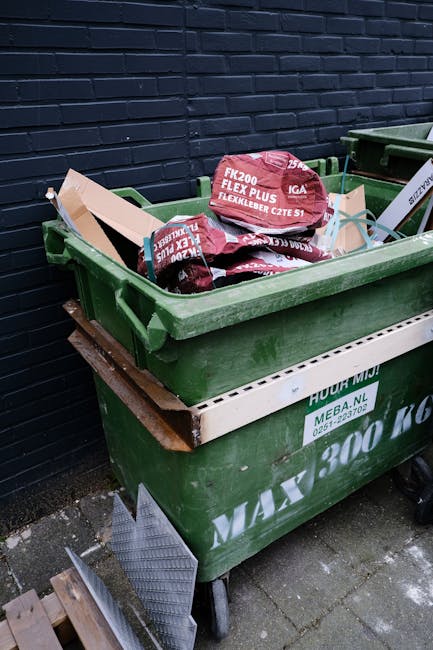 A waste collection worker wearing a high-visibility yellow vest and dark clothing is operating a large urban rubbish collection vehicle on a city street during early evening or dusk. The vehicle has r