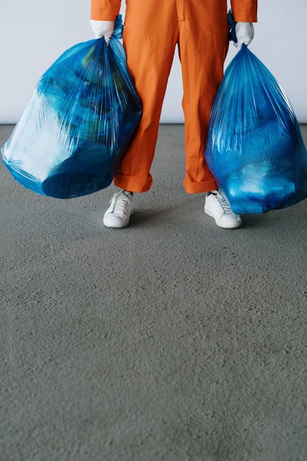 A person standing on a grey indoor floor, holding two large blue plastic rubbish bags, one in each hand. The individual is dressed in orange work overalls and white sneakers, with only their lower bod