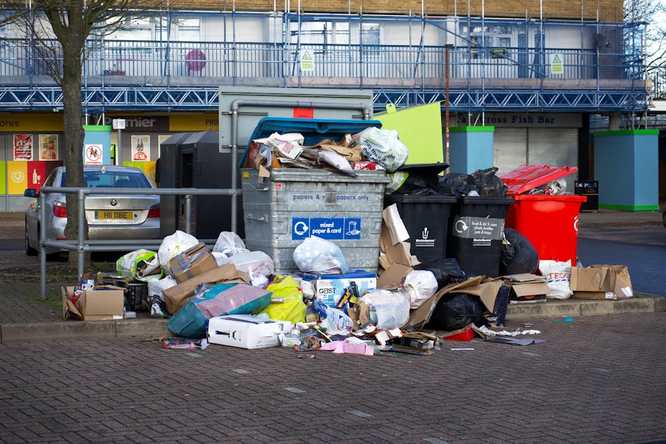 A large collection of mixed waste and rubbish overflow from open-top dumpsters placed on a paved sidewalk in an urban area. The waste includes crumpled cardboard boxes, plastic shopping bags, paper pa