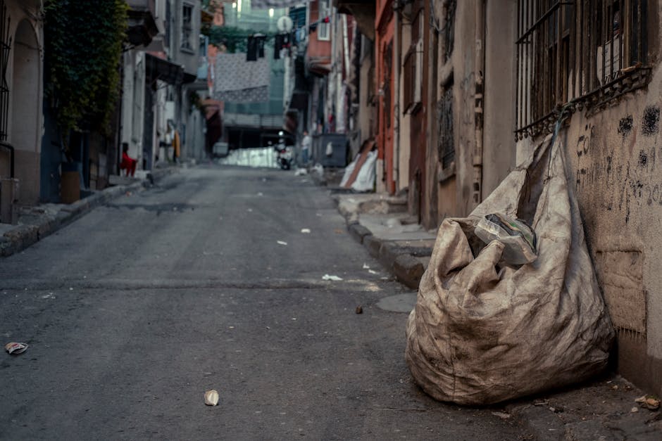 A large collection of mixed waste and rubbish overflow from open-top dumpsters placed on a paved sidewalk in an urban area. The waste includes crumpled cardboard boxes, plastic shopping bags, paper pa